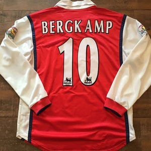 May include: Red and white long-sleeved soccer jersey with the name "BERGKAMP" and the number "10" on the back. The sleeves are white with blue trim, and the jersey has a white collar. Logos are visible on the sleeves and below the number.