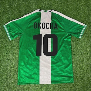 May include: Green soccer jersey with white accents and the name "OKOCHA" and the number "10" in black. The jersey has a white stripe down the center and white stripes on the sleeves. The jersey is made of a shiny material.