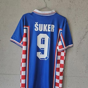 May include: Blue soccer jersey with the name "ŠUKER" and the number "9" on the back. The jersey has white and red checkered accents on the sides and a white collar with blue and red trim. The jersey is hanging on a metal hanger.