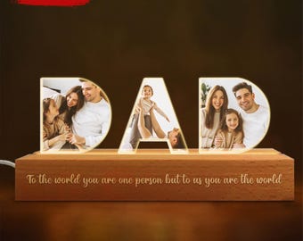 Könnte beinhalten: Eine Holz-LED-Leuchte mit dem beleuchteten Wort "DAD". Jeder Buchstabe zeigt ein Familienfoto. Auf der Basis steht der Text "To the world you are one person but to us you are the world". Oben links steht "DESIGNS CAN BE CHANGED".
