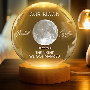 Puede incluir: Una esfera de cristal iluminada sobre una base de madera, con una imagen detallada de la luna. La esfera está inscrita con "OUR MOON", los nombres "Michael" y "Sophia", la fecha "21.02.2019" y el texto "THE NIGHT WE GOT MARRIED."