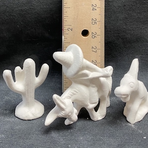 Puede incluir: Tres figuritas de cerámica blanca: un cactus, un burro con una persona montándolo y un burro. El burro con el jinete lleva un sombrero.
