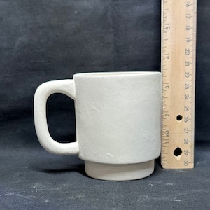 Puede incluir: Una taza de cerámica blanca con asa. La taza mide aproximadamente 10 cm de alto y tiene un diseño sencillo.