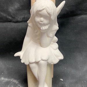 Puede incluir: Figura de hada de cerámica blanca, con expresión pensativa, apoyando la barbilla en la mano. Viste una falda con volantes y tiene orejas puntiagudas. La figura mide aproximadamente 15 cm de altura.