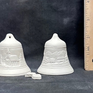 May include: Two white ceramic Christmas bells with a winter scene design. The bell on the left has a church and trees, while the bell on the right has a mountain scene. The bells are approximately 3 inches tall.