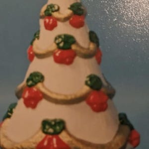 Può includere: Un ornamento per albero di Natale in ceramica bianca con una stella dorata in cima. L'albero è decorato con ornamenti rossi e verdi e un nastro dorato.