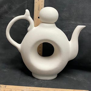 May include: White ceramic teapot with a round hole in the centre and a round knob on top. The teapot is approximately 25 cm tall.