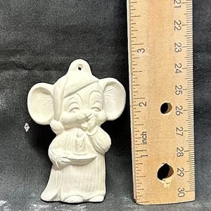 May include: A ceramic mouse figurine, painted white, with large ears and a smiling face. The mouse is wearing a hat and holding a plate. A wooden ruler is next to the mouse, with inch measurements.