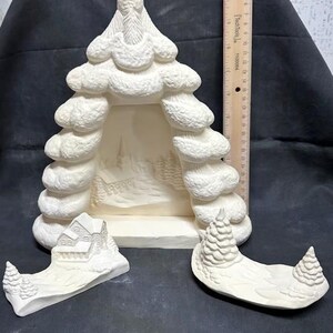 May include: A white ceramic Christmas tree with a cutout in the center, a separate piece with a house and a separate piece with two trees. The tree is decorated with a snowy texture.