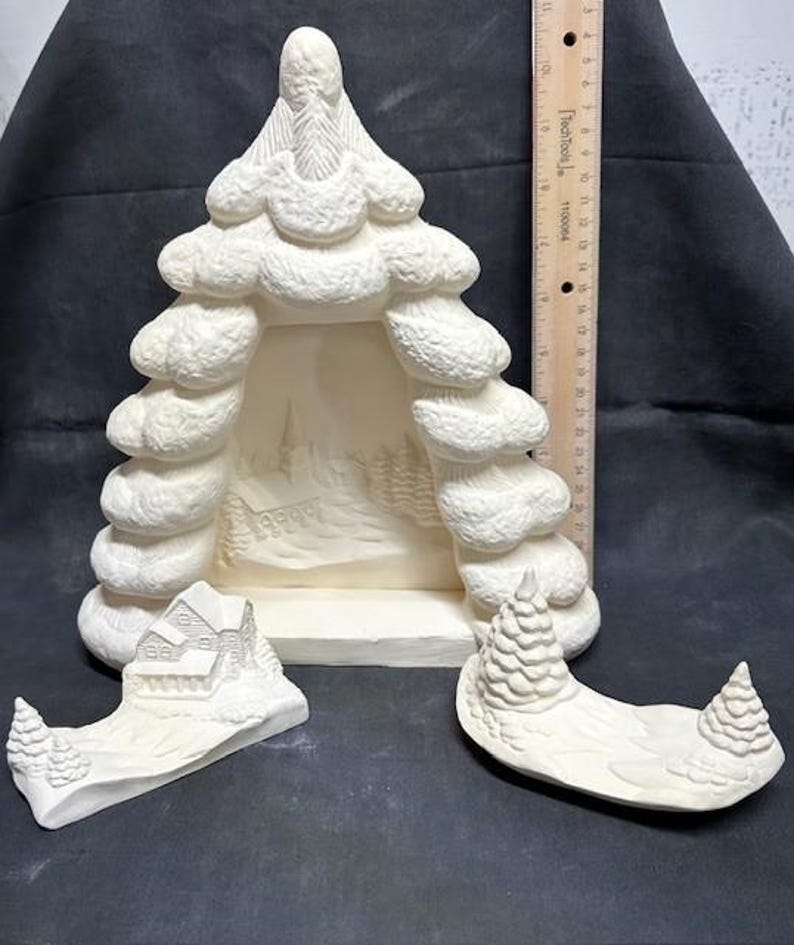 May include: A white ceramic Christmas tree with a cutout in the center, a separate piece with a house and a separate piece with two trees. The tree is decorated with a snowy texture.