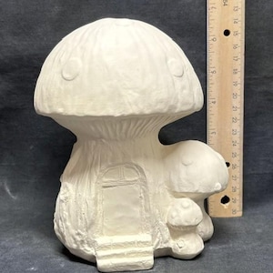 Può includere: Una statuina a forma di casa a fungo in ceramica bianca. Il fungo ha un grande cappello con tre piccoli cerchi e una porta con gradini. Un fungo più piccolo è attaccato al lato. La statuina è alta circa 25 cm.