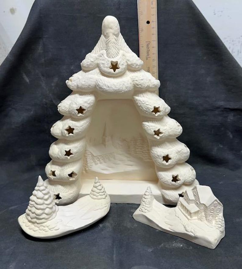 May include: A white ceramic Christmas tree with cut-out stars and a winter scene inside. The tree is in three pieces, with a base, a middle section, and a top. The base has a small house and two trees, while the middle section has a winter scene with trees and a snowy path. The top of the tree has a star on top.