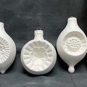 May include: Three white ceramic Christmas ornaments with embossed floral designs. The ornaments are teardrop-shaped and have a small metal loop for hanging. A wooden ruler is next to the ornaments, showing measurements in inches.