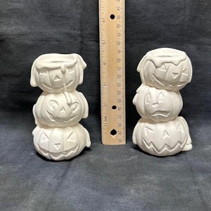 Puede incluir: Dos juegos de tres portavelas de cerámica blanca en forma de calabaza. Cada juego está apilado con una calabaza de diferente tamaño, con la calabaza más pequeña en la parte inferior y la más grande en la parte superior. Las calabazas tienen caras de calabaza tallada.