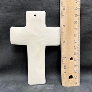 May include: A plain, off-white ceramic cross with a hole at the top for hanging. The cross is approximately 5 inches tall. A wooden ruler is placed next to the cross for scale, showing inch measurements.
