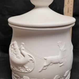 Puede incluir: Tarro de galletas de cerámica blanca con tapa. El tarro presenta un diseño en relieve de Papá Noel en su trineo tirado por renos. La parte inferior del tarro tiene un diseño en relieve de casas.