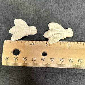 Könnte beinhalten: Zwei kleine, unbemalte Bienenfiguren. Die Bienen befinden sich in der Nähe eines Holzlineals und sind jeweils etwa 2,5 cm lang. Die Flügel und Körperdetails sind sichtbar. Der Hintergrund ist ein dunkelgrauer Stoff.