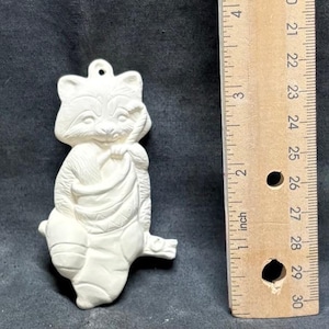 May include: A ceramic raccoon figurine, unpainted, with a loop for hanging. The raccoon is holding a sack and measures approximately 10 cm in height. The figurine is a light cream colour and has detailed facial features.