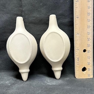 Può includere: Due pezzi in ceramica bianco sporco a forma di goccia, con un'indentazione ovale e un fondo appuntito. Un righello di legno con misure in pollici è accanto ai pezzi.