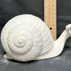 Puede incluir: Figurita de caracol de cerámica blanca con una concha detallada y un cuerpo liso. El caracol mide aproximadamente 15 cm de largo.