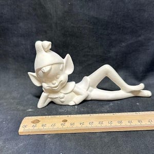 Puede incluir: Figurilla de cerámica blanca de un duende de Navidad acostado de lado con los brazos cruzados. El duende lleva un gorro puntiagudo y tiene una expresión traviesa en su rostro.