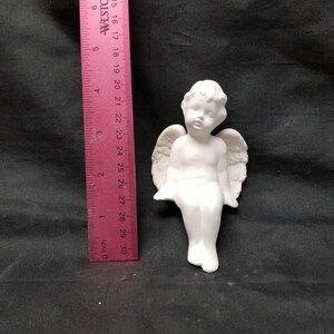 May include: White ceramic angel figurine with wings, sitting with legs crossed. The angel has a cherubic face and is approximately 6 inches tall.