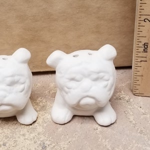 May include: Two white ceramic bulldog salt and pepper shakers. Each bulldog has a detailed face with a furrowed brow and small holes on top. A wooden ruler is next to the shakers.