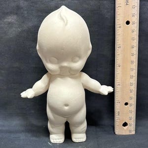 Könnte beinhalten: Unbemalte Kewpie-Puppenfigur aus Biskuitporzellan, bereit zur individuellen Gestaltung. Die Puppe hat einen runden Kopf mit einer kleinen Locke, ausgestreckte Arme und einen prallen Bauch. Ein Lineal liegt neben der Puppe mit Zentimetermessungen.