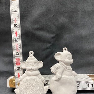 以下が含まれることがあります： マウスの形をした白いセラミック製のクリスマスオーナメントが2つ。1つのマウスは丸い物体を持ち、もう1つはスカーフをしています。