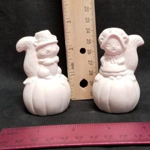 May include: Two white ceramic figurines of squirrels sitting on pumpkins. The squirrel on the left is wearing a pilgrim hat and the squirrel on the right is wearing a bonnet.