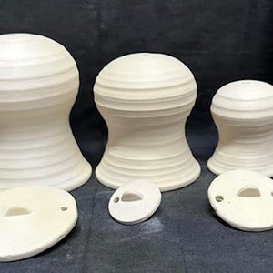 May include: A collection of three off-white ceramic pieces, each with a rounded top and a flared base. Two matching oval lids are also present. A wooden ruler is in the background.