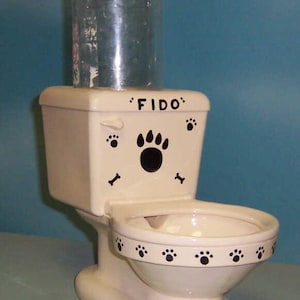 Può includere: Un abbeveratoio per cani a forma di toilette, color crema, con impronte di zampe nere e il nome "FIDO". Una bottiglia di plastica trasparente è attaccata sopra. Un accessorio divertente per animali domestici.