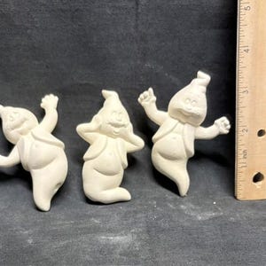 May include: Three unpainted ceramic ghost figurines with outstretched arms and smiling faces. Each ghost is wearing a vest. A wooden ruler with inch measurements is next to the ghosts.