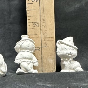 May include: Four unpainted Halloween figurines: a smiling jack-o'-lantern, a scarecrow, a cat wearing a witch hat, and a baby on a pumpkin. A wooden ruler with inch markings is in the background.