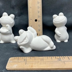 Könnte beinhalten: Drei weiße Keramik-Froschfiguren. Ein Frosch steht aufrecht, ein anderer liegt auf dem Rücken und der dritte steht mit verschränkten Armen. Die Frösche sind alle weiß bemalt und haben schwarze Augen.