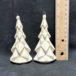 Puede incluir: Dos adornos navideños geométricos en forma de árbol, de color blanquecino. Cada árbol tiene un diseño facetado y un lazo de alambre para colgar. Los árboles miden unos 10 cm de alto. Fondo oscuro.