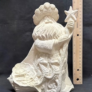 Puede incluir: Escultura de cerámica blanquecina de Papá Noel con barba y sombrero largos, sosteniendo una varita de estrella de mar. La escultura presenta detalles de conchas marinas e hipocampos, y mide aproximadamente 25 cm de altura.