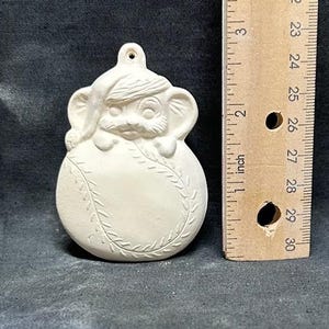 May include: Unpainted ceramic ornament in the shape of a baseball with a mouse wearing a Santa hat. The ornament is approximately 3 inches tall and has a loop for hanging. A ruler is next to the ornament.