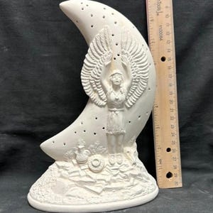 Op de afbeelding: Witte keramische halve maansculptuur met een vrouw met vleugels die in het midden staat. De vrouw houdt haar armen omhoog en draagt een kralenketting. De maan heeft gaten en een getextureerde basis.