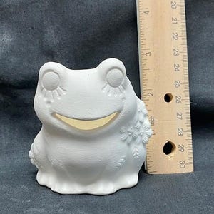 Könnte beinhalten: Weiße Keramik-Froschfigur mit einem lächelnden Gesicht und geschlossenen Augen. Der Frosch hat ein Blumenmuster an der Seite und ist etwa 7,5 cm hoch.