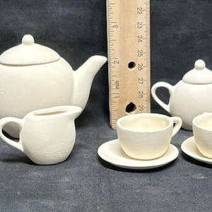 May include: A white ceramic tea set, including a teapot, creamer, sugar bowl, and two teacups with saucers. The teapot is approximately 6 inches tall. The set is unpainted and ready for customization.