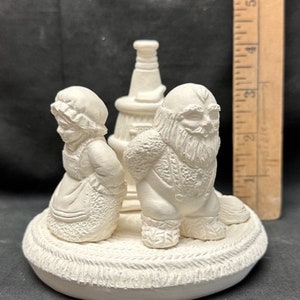 May include: White ceramic figurine set of a woman and a man with a beard, standing next to a tall, narrow structure. The figurines are on a round base with a rope-like design.