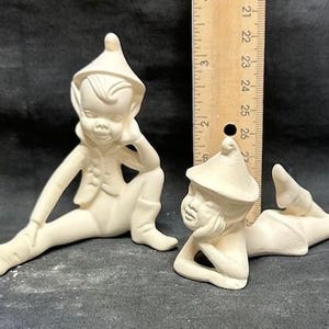 May include: Two unpainted ceramic elf figurines. One is seated with a leg extended, the other is lying down. Both have pointed hats and are in a relaxed pose. A wooden ruler is placed between them.