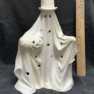 Könnte beinhalten: Eine weiße Keramik-Geisterfigur mit Zylinder und geschnitztem Kürbis. Das drapierte Tuch des Geistes hat sternförmige Aussparungen. Eine Halloween-Dekoration, die jedem Zuhause eine gruselige Note verleiht.