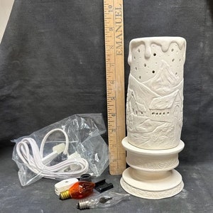 Puede incluir: Portavelas de cerámica blanca con un diseño de escena de invierno. El portavelas tiene forma de vela con una base y una parte superior extraíble. La escena incluye una montaña nevada, una pequeña casa y árboles. El portavelas viene con un cable, una bombilla y un interruptor.