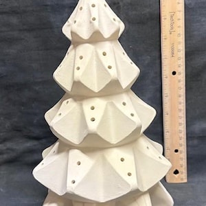 Può includere: Un albero di Natale in ceramica bianca con base a forma di stella e design geometrico. L'albero ha più strati con piccoli fori distanziati. Un righello di legno è posto accanto all'albero.