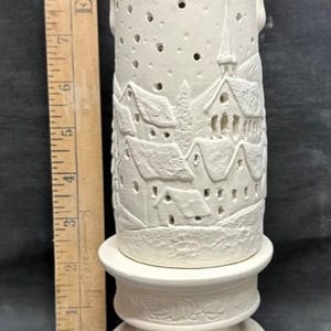 Könnte beinhalten: Ein weißer Keramik-Kerzenhalter mit einer eingeschnitzten Winterdorf-Szene. Der zylindrische Halter hat kleine Löcher und steht auf einem dekorativen Sockel. Der Halter ist etwa 15 cm hoch.