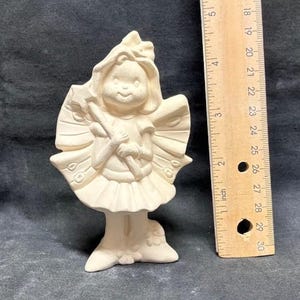 May include: A bisque ceramic fairy figurine with a smiling face, holding a wand. The fairy has a ruffled skirt and detailed wings. A wooden ruler is next to the figurine, measuring up to 30 inches.