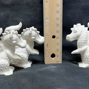 May include: Three white ceramic dragon figurines, two are sitting side-by-side and one is sitting separately. The dragons have wings and tails.