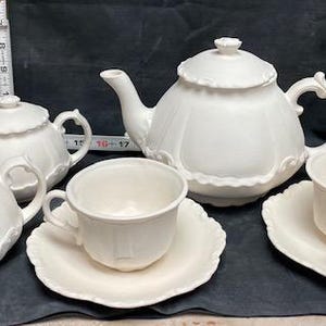 Op de afbeelding: Een wit porseleinen theeservies, inclusief een theepot, suikerpot, melkkan en twee kopjes met schotels. De set heeft een geschulpte rand. Een meetlint staat op de achtergrond.